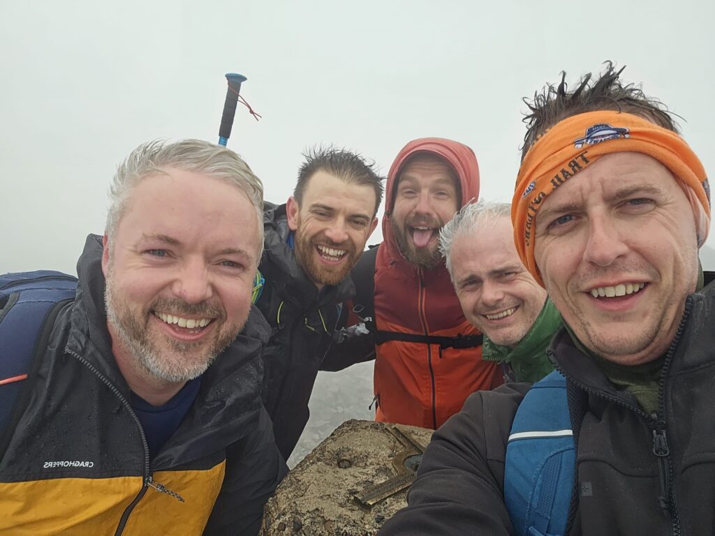 A group of five men smile huddled together as they stand atop of a mountain.