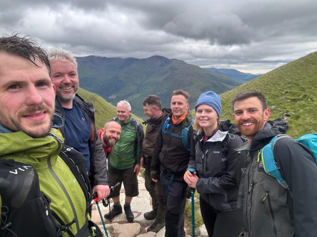 A group of 7 men and a woman smile huddled together as they stand atop of a mountain.