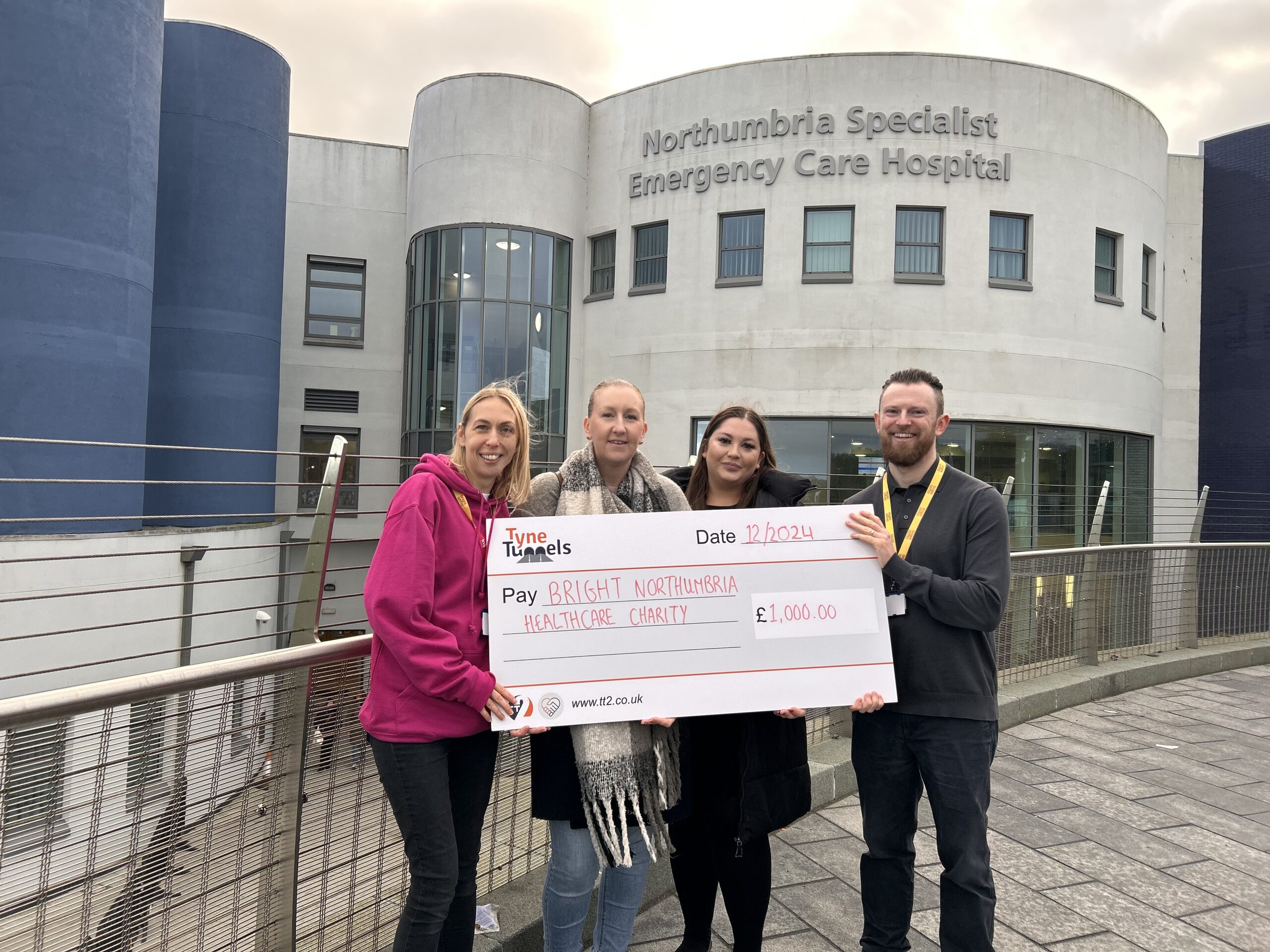 Two Bright team members hold a giant cheque, presented to them by two female employees of the Tyne Tunnel. The cheque is for £1,000.