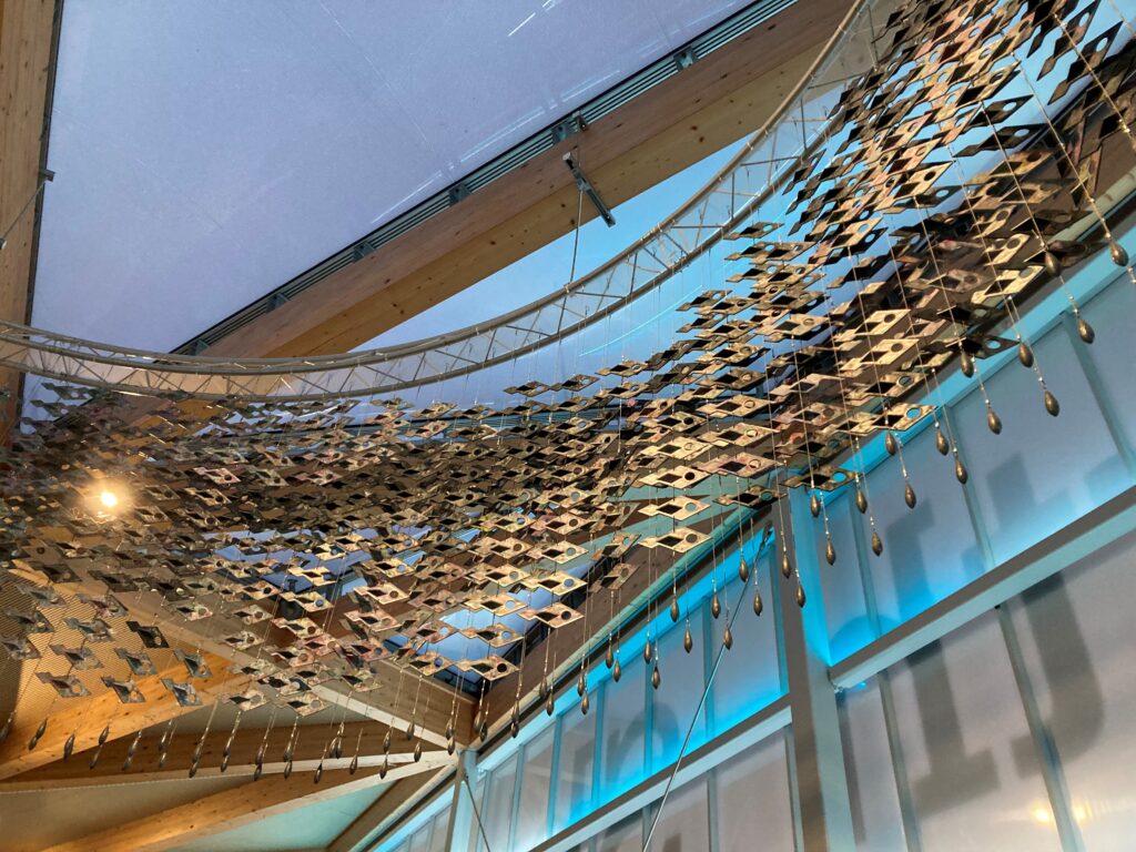 A large-scale suspended art installation featuring numerous metallic fish-shaped pieces connected in a flowing, net-like formation. Each fish has a circular cutout, and they are arranged in a wave-like pattern, evoking the movement of a shimmering school of fish. Small teardrop-shaped weights hang from the lower edges, enhancing the sense of fluidity. The installation is suspended from a curved metal frame attached to the ceiling of a modern space with wooden beams and glass walls, illuminated by soft blue lighting.