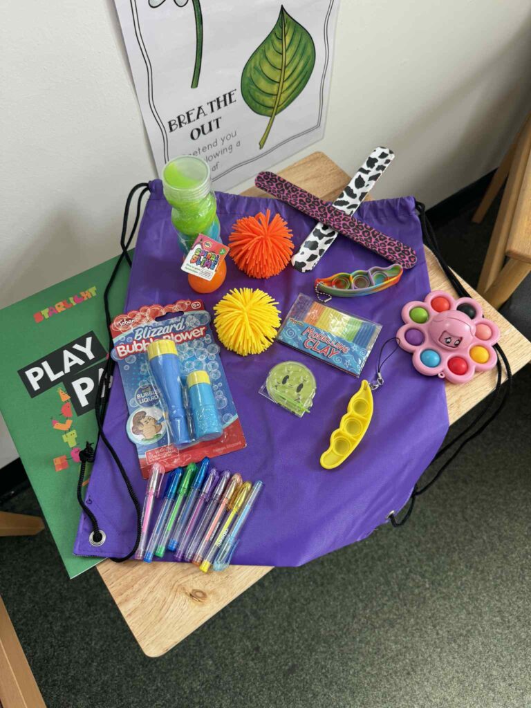 A purple drawstring bag sits flat on top of a small wooden table. Across the bag, various fidget toys are shown - including a bubble blower, stress balls, and pop its.