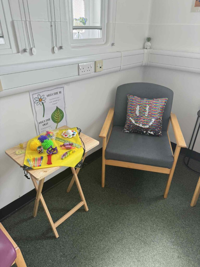 This picture shows more of the room than the previous one. The small table and bag are there, with a grey armchair to the right of it. On the armchair, there is a sequinned pillow with a smiley face drawn on it.